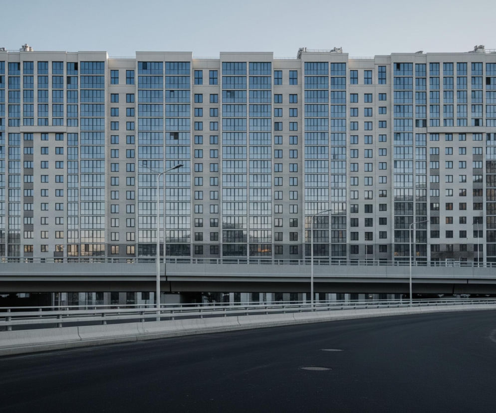 Wysoki, nowoczesny budynek mieszkalny o jasnej elewacji dominuje w górnej części obrazu, rozciągając się horyzontalnie przez niemal całą jego szerokość. Fasada budynku składa się z regularnie rozmieszczonych okien, z których wiele ma odbicia niebieskiego nieba, oraz z mniejszych, prostokątnych otworów w szarych ścianach. Poniżej budynku biegnie estakada, widoczna jako szeroki, betonowy pas z metalowymi barierkami, a pod nią ciemny przejazd. Na pierwszym planie znajduje się gładka, czarna nawierzchnia drogi, która zakręca w prawo, ograniczona betonową barierą i metalowymi słupami latarni ulicznych, oświetlających drogę i estakadę.