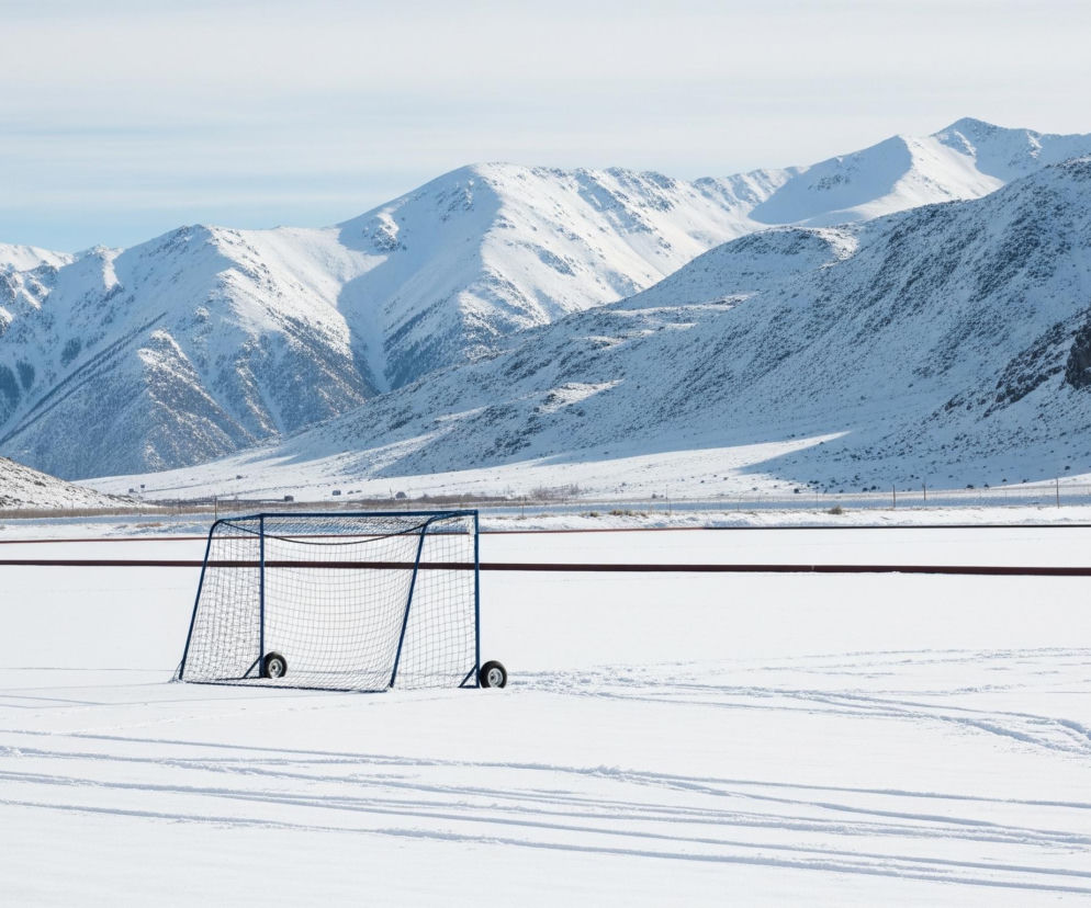 Na ośnieżonym boisku w centrum kadru stoi niebieska bramka z siatką, wyposażona w koła u podstawy. Za bramką rozciąga się płaskie, białe pole, zakończone czerwoną linią, a dalej widoczne są pagórki pokryte resztkami śniegu. W tle dominują majestatyczne, ośnieżone szczyty górskie, a nad nimi rozpościera się jasne, lekko zachmurzone niebo. Na pierwszym planie, w białym śniegu, widać liczne ślady przypominające tory sanek lub nart.