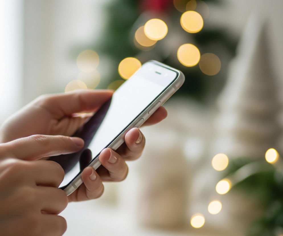 Kobieta trzyma smartfon w obu dłoniach, lewa dłoń dotyka ekranu kciukiem. Telefon ma biały ekran i srebrne krawędzie, a na paznokciach kobiety widoczny jest jasny lakier. W tle, rozmyte, znajdują się żółte lampki choinkowe oraz fragmenty zielonego drzewka i czerwonych ozdób.