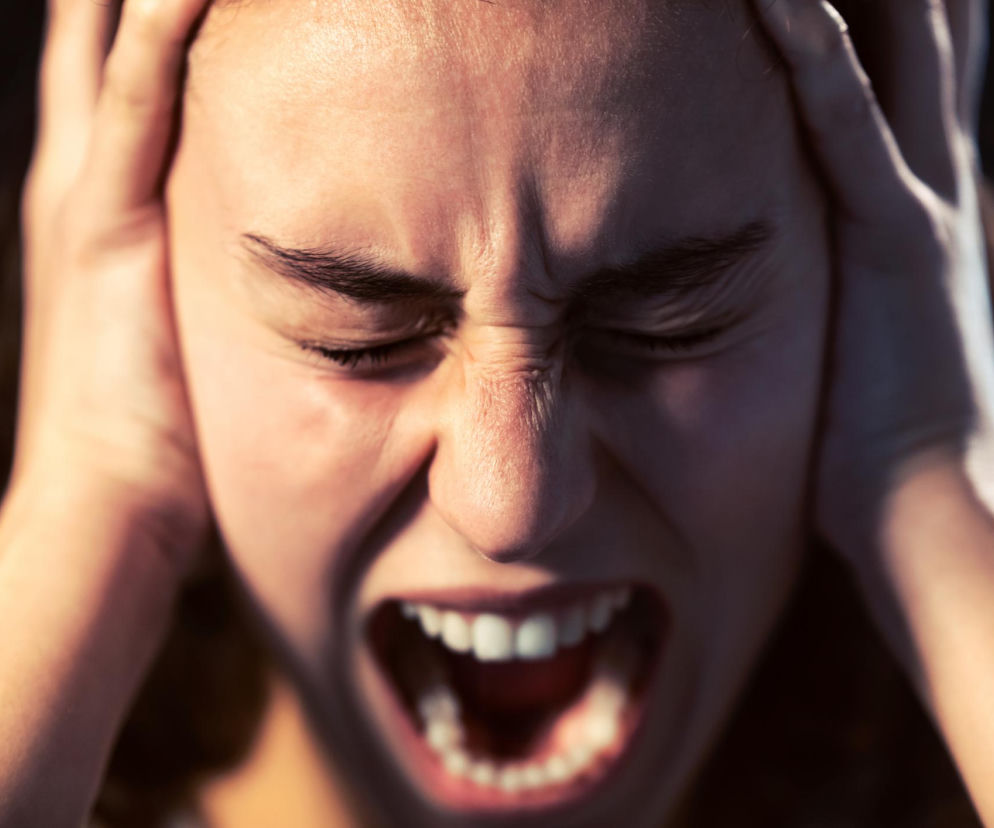 Kobieta w stresie z zamkniętymi oczami i otwartymi ustami, krzycząca z frustracji. Dłonie przyłożone do skroni, symbolizują chroniczny stres i przepracowanie, które negatywnie wpływają na zdrowie mózgu. Dowiedz się więcej o szkodliwych nawykach na Poradnik Zdrowie.
