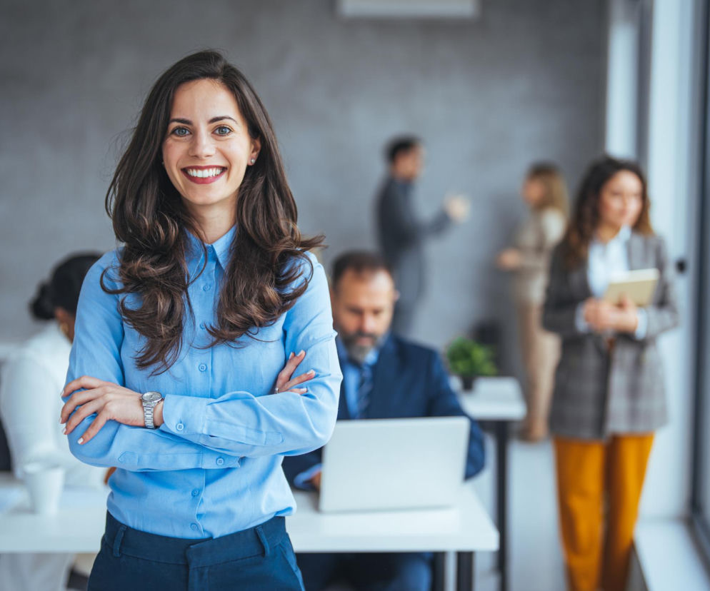 Uśmiechnięta kobieta z długimi, ciemnymi włosami w błękitnej koszuli i granatowych spodniach, ze skrzyżowanymi rękami. W tle, w rozmazanym biurze, inni pracownicy i biurka. Symbolizuje rozwój przedsiębiorczości w Polsce, o czym można przeczytać na Super Biznes.