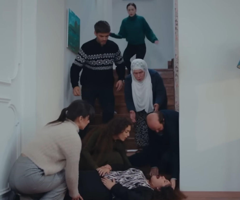 Kobieta leżąca na podłodze u podnóża schodów, otoczona przez zaniepokojonych ludzi, w tym starszą kobietę w hidżabie, młodą kobietę w zielonym golfie i mężczyznę w swetrze. Scena z serialu Dziedzictwo, gdzie Cansel symuluje upadek ze schodów po poronieniu, oskarżając Nanę, o czym przeczytasz na Super Seriale.