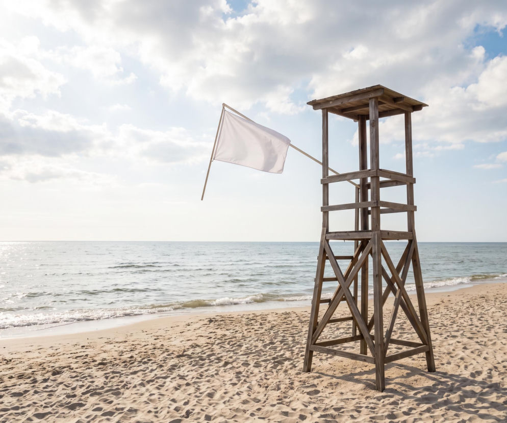 Drewniana wieża ratownika z białą flagą na plaży w Kołobrzegu. O czystej wodzie przeczytasz na SE Szczecin.