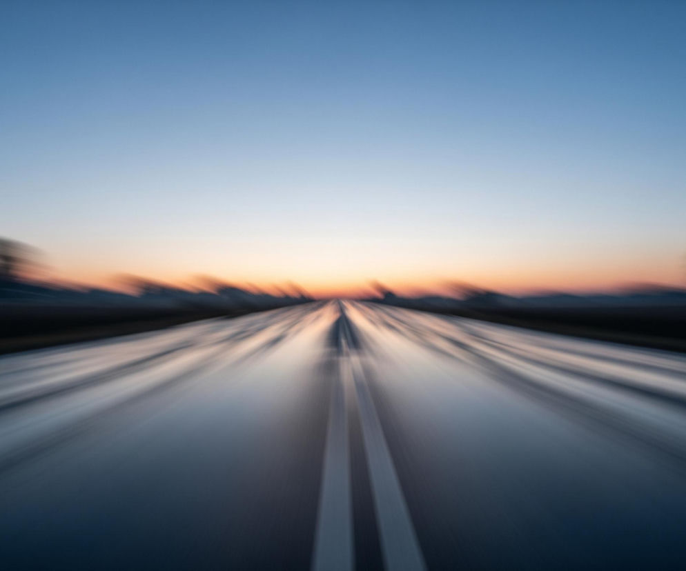 Asfaltowa droga rozciąga się w głąb obrazu, prowadząc wzrok ku horyzontowi. Na niej znajdują się dwie białe, przerywane linie, zbiegające się w oddali. Perspektywa zniekształcona jest efektem rozmycia ruchu, co nadaje wrażenie dużej prędkości. W oddali niebo przechodzi od jasnego błękitu u góry, poprzez jaśniejszy odcień błękitu, do pomarańczowo-różowego paska, który jest najjaśniejszym elementem obrazu, sugerującym wschód lub zachód słońca. Po bokach drogi, tuż nad horyzontem, widoczne są rozmyte, ciemne sylwetki drzew lub krajobrazu, również zniekształcone przez efekt ruchu.