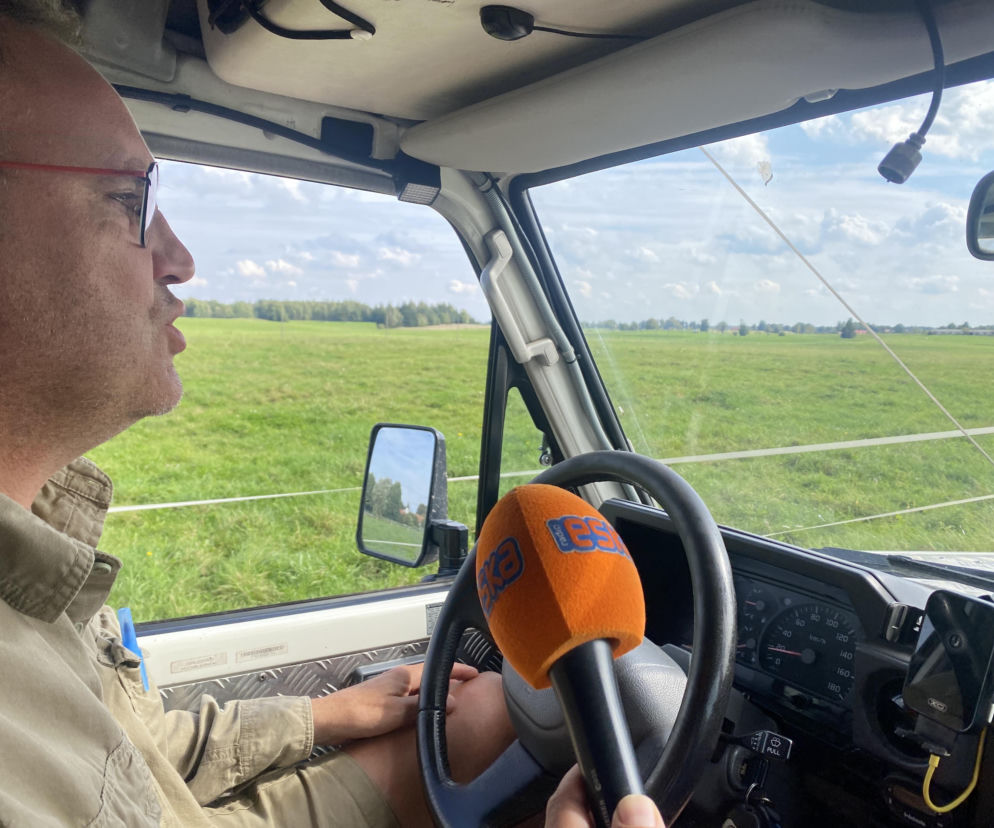 Dziennikarz z mikrofonem Eska Olsztyn w aucie na tle Polany Ukciańskiej. O proteście mieszkańców przeczytasz na Eska Olsztyn.