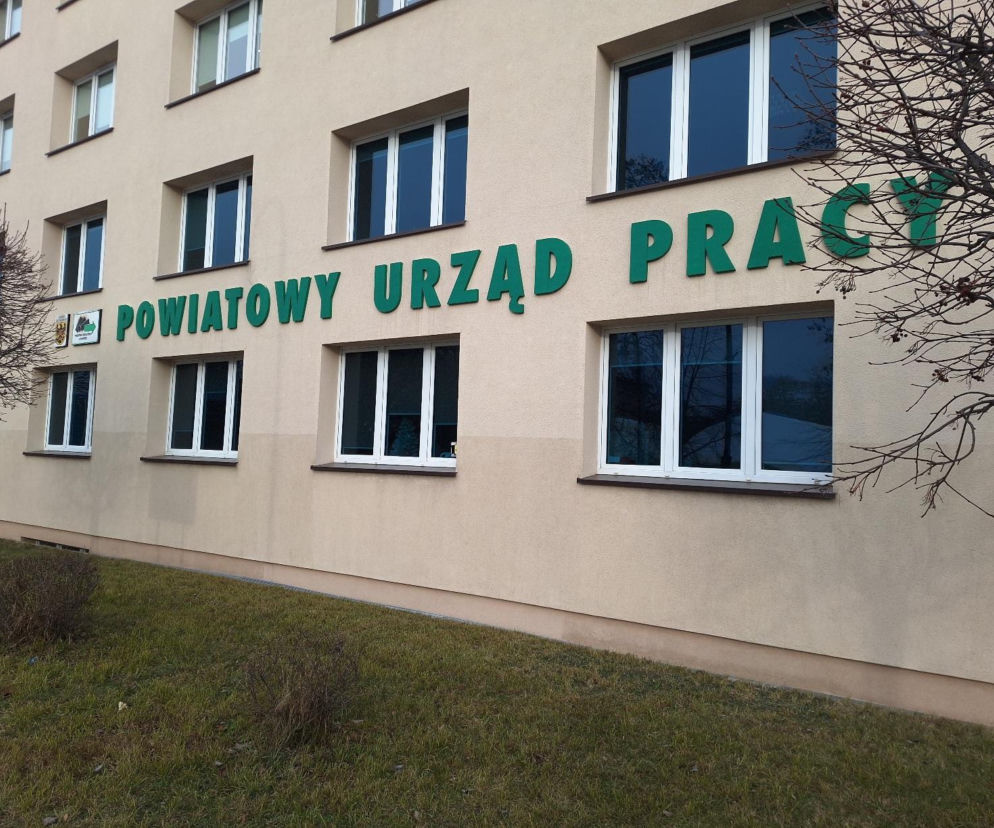 Budynek Powiatowego Urzędu Pracy w Głogowie z zielonym napisem i herbem na beżowej fasadzie, otoczony trawnikiem i nagimi drzewami. Odnosi się do artykułu o wzroście bezrobocia w Polsce, o czym przeczytasz na Super Biznes.