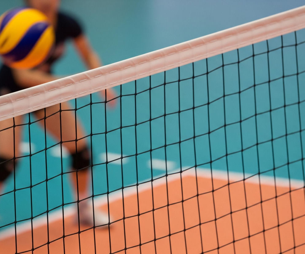 Siatka do siatkówki, z białym obramowaniem i czarnymi oczkami, zajmuje większą część kadru. Za siatką widoczna jest rozmyta postać osoby w ciemnym stroju sportowym, przygotowującej się do odbicia żółto-niebieskiej piłki do siatkówki. Tło stanowi jasnoniebieska ściana i pomarańczowy fragment podłogi boiska, a po lewej stronie siatki widać fragment słupka w czerwono-białe pasy.