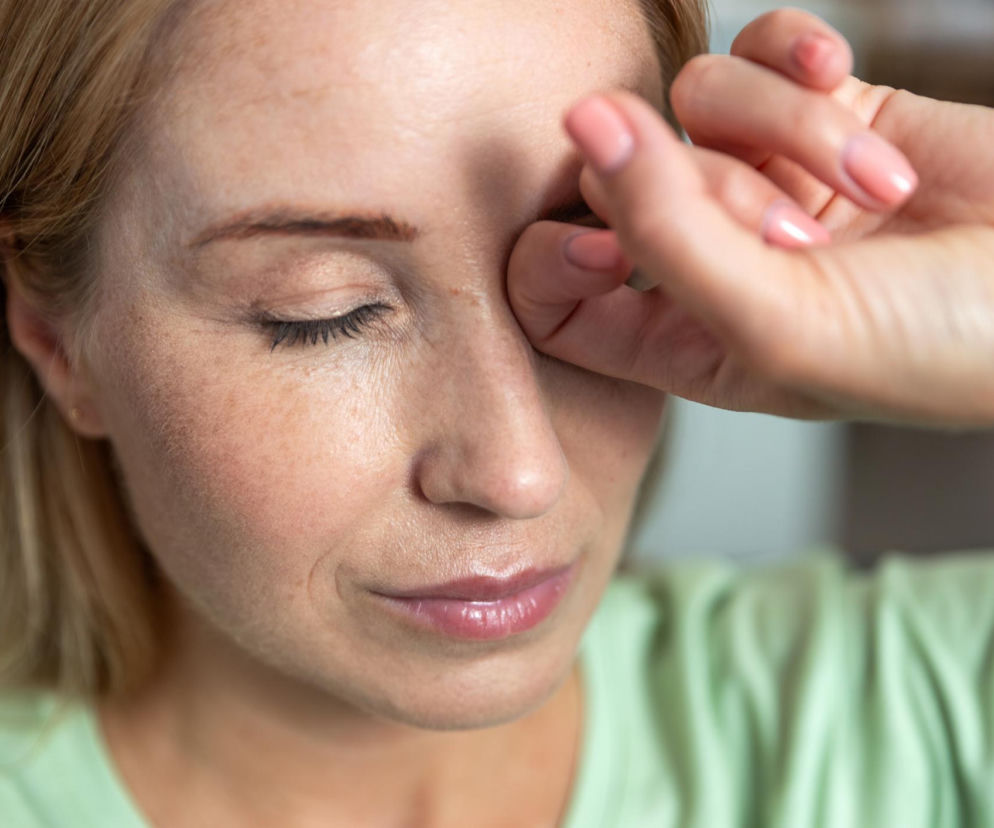 Kobieta z blond włosami w zielonej koszulce, z zamkniętymi oczami, masuje prawą powiekę palcami z różowymi paznokciami. Zdjęcie ilustruje problem drgającej powieki i odnosi się do artykułu na Poradnik Zdrowie o tym, kiedy skacząca powieka to nie tylko brak magnezu.
