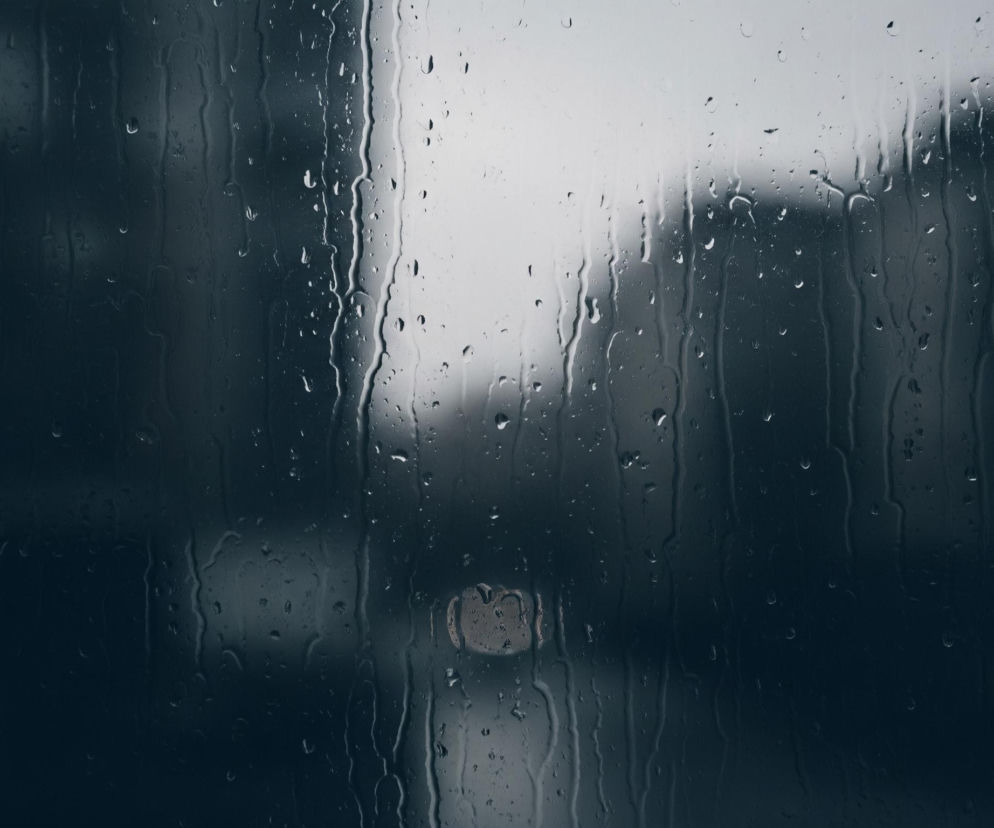 Przednia szyba samochodu pokryta jest licznymi kroplami wody, z których niektóre spływają, tworząc smugi. W tle, rozmyte i niewyraźne, widoczne są kontury budynków w ciemnoszarym kolorze, a na górze i po prawej stronie jaśniejsze, zamglone niebo. W centralnej części obrazu, na dole, widoczny jest okrągły, jaśniejszy kształt, prawdopodobnie odległe światło. Dominują ciemne, chłodne barwy, tworząc melancholijny nastrój.