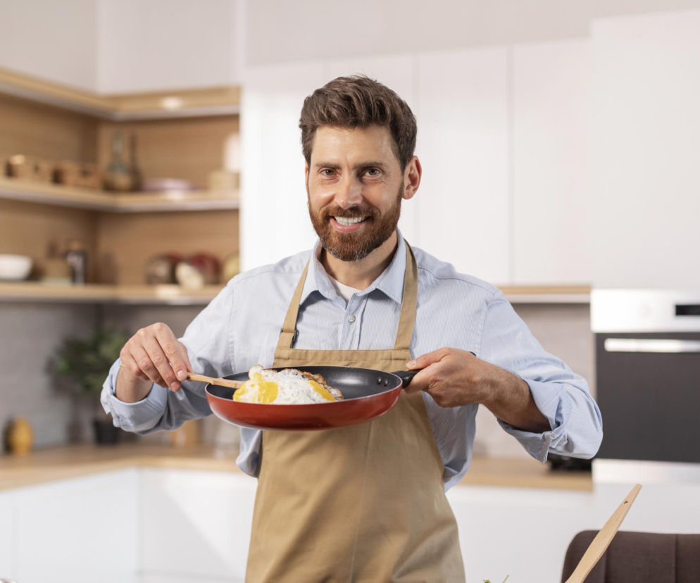 Uśmiechnięty mężczyzna w jasnej koszuli i beżowym fartuchu trzyma patelnię z jajkami sadzonymi. W tle nowoczesna kuchnia z drewnianymi półkami. Na Poradnik Zdrowie znajdziesz więcej o wpływie jajek na mózg.