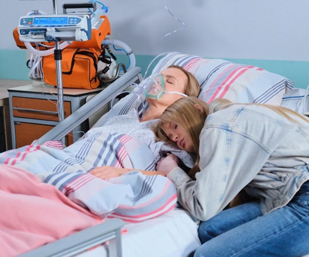 Kobieta w masce tlenowej leży na szpitalnym łóżku, a młoda dziewczyna śpi przytulona do jej boku. Scena z serialu „Pierwsza miłość”, w której Marysia (Aneta Zając) walczy o życie po wypadku. Więcej dramatycznych wątków znajdziesz na Super Seriale.