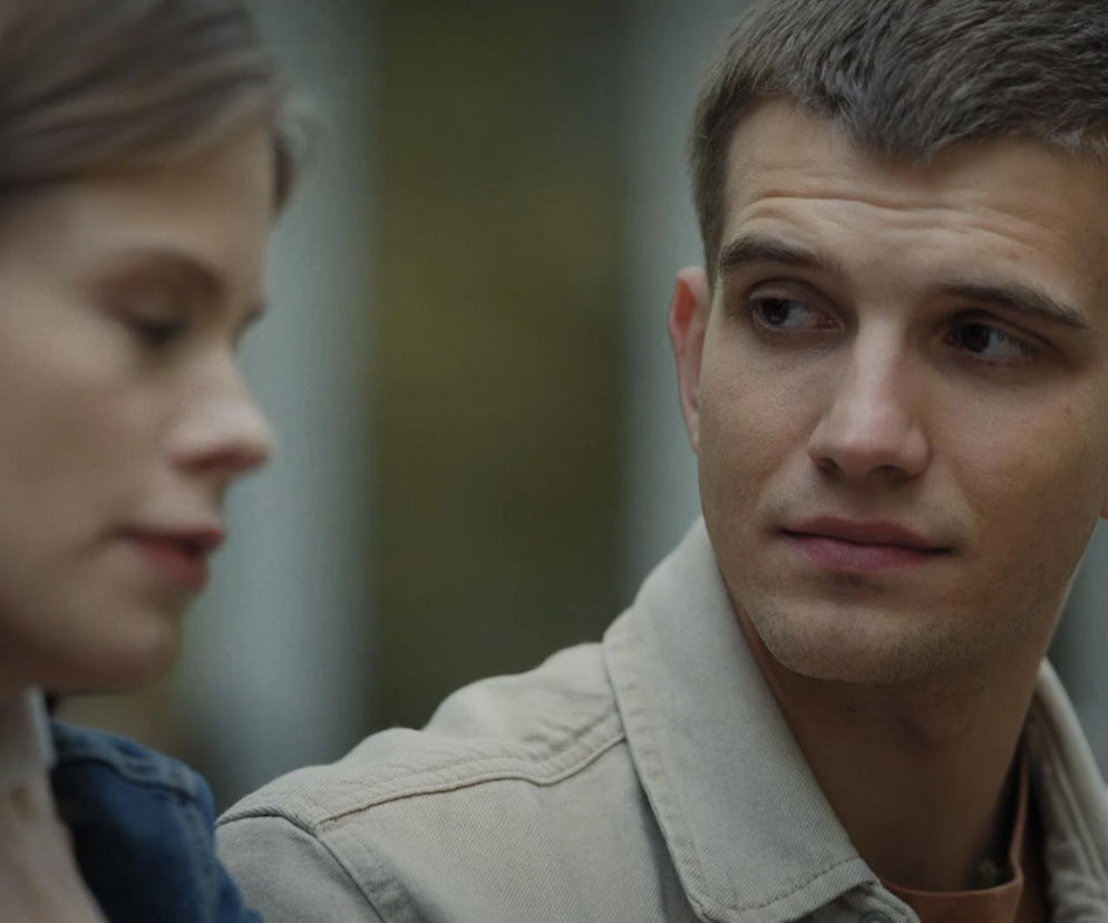 Mateusz (Rafał Kowalski) i Majka (Matylda Giegżno) z serialu M jak miłość. Mateusz, ubrany w beżową koszulę, spogląda w prawo z zaniepokojeniem, podczas gdy Majka, ubrana w dżinsową kurtkę, patrzy na niego. Kontekst odcinka 1922 sugeruje ich skomplikowaną relację, o czym przeczytasz więcej na portalu Super Seriale.