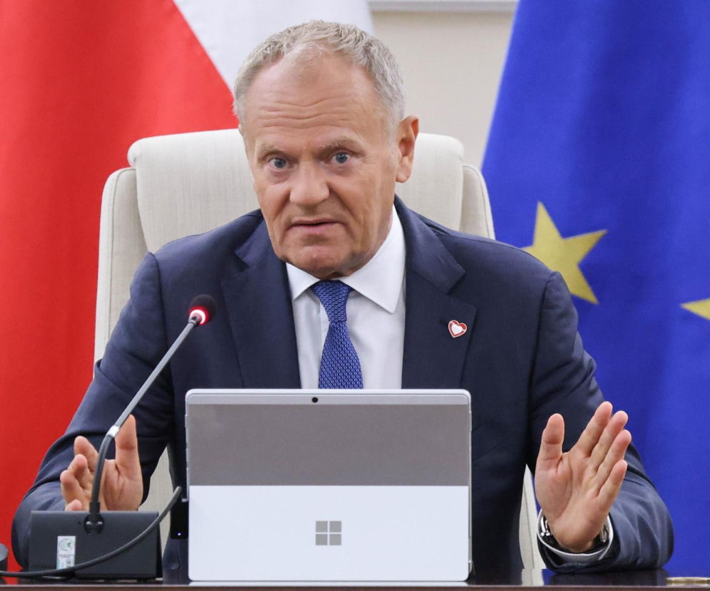 Premier Donald Tusk przy stole z laptopem na tle flag Polski i Unii Europejskiej. O budżecie na 2027 rok przeczytasz w Esce.