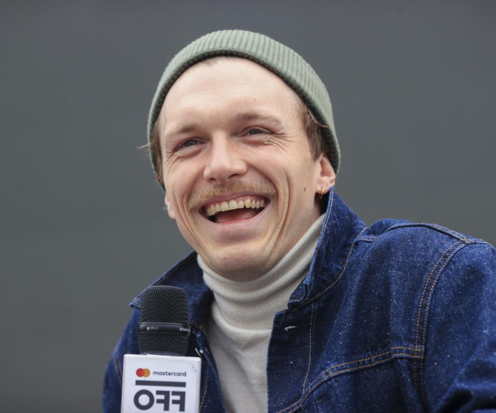 Uśmiechnięty aktor Tomasz Włosok w zielonej czapce i jeansowej kurtce. O metamorfozach gwiazdy przeczytasz na SE.