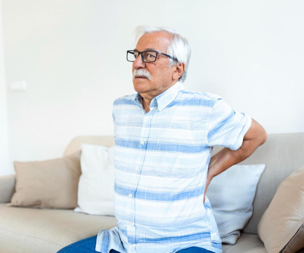 Starszy mężczyzna w okularach i koszuli w paski trzyma się za bolące plecy, siedząc na kanapie z jasnymi poduszkami. Obraz symbolizuje problemy ze stawami i brak ruchu, które mogą prowadzić do bólu. Dowiedz się więcej o zdrowych stawach na portalu Poradnik Zdrowie.