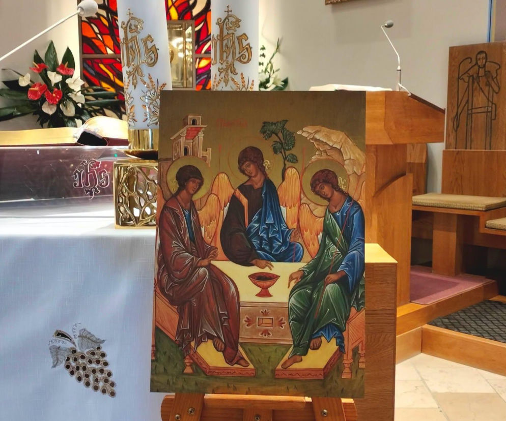 Ikona Trójcy Świętej Andrieja Rublowa, symbol gościnności i wspólnoty parafialnej, ustawiona na drewnianym stojaku w kościele. Obok niej widoczne są zapalone świece ołtarzowe, zdobiony mszał i elementy wystroju świątyni. Więcej o życiu parafii przeczytasz na naszym portalu.