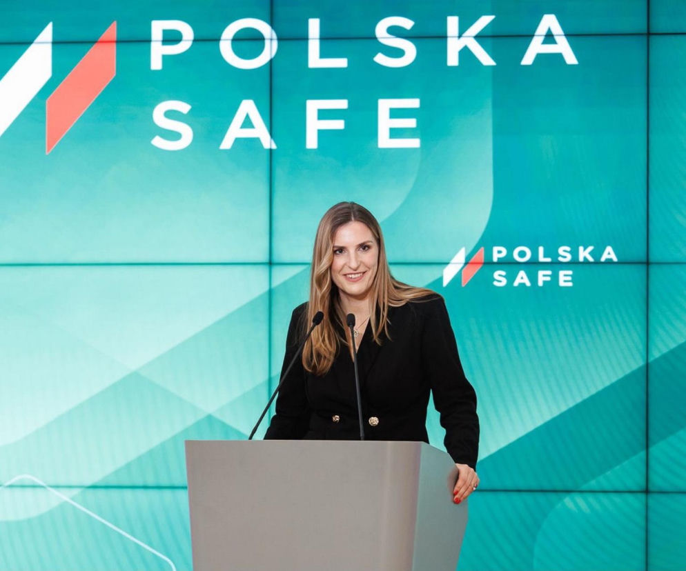Magdalena Sobkowiak-Czarnecka, pełnomocniczka rządu ds. programu SAFE, uśmiechnięta stoi za mównicą, przemawiając na tle turkusowego ekranu z napisem POLSKA SAFE. Widoczne mikrofony na mównicy. O kluczowej roli programu SAFE dla polskiej zbrojeniówki przeczytasz na naszym portalu.