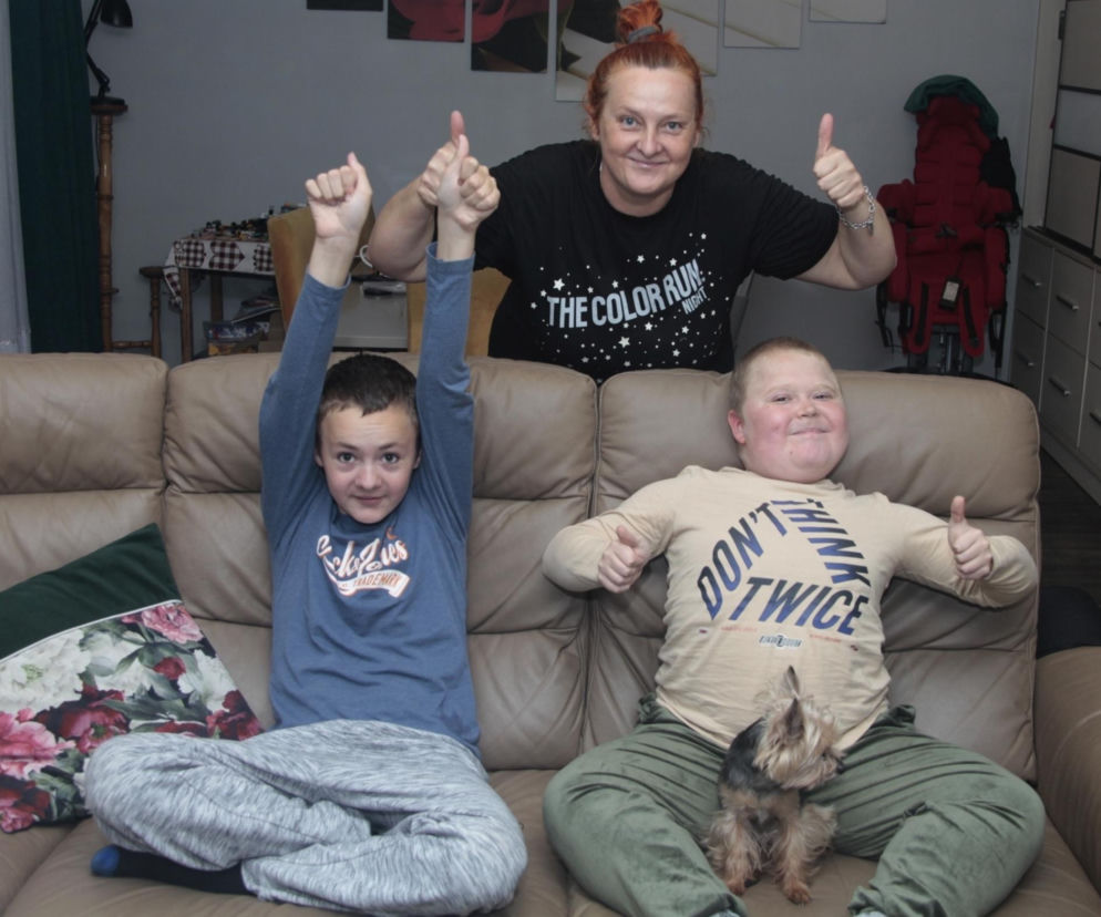 Tymoteusz Kalata, jego brat i mama Renata Kalata uśmiechają się do zdjęcia, siedząc na kanapie i pokazując kciuki w górę. Tymek, w beżowej bluzce, leży z małym psem, a jego brat siedzi obok, podnosząc ręce. Rodzina wyraża radość z sukcesu najdroższej kuracji. Więcej o ich historii przeczytasz na naszym portalu.