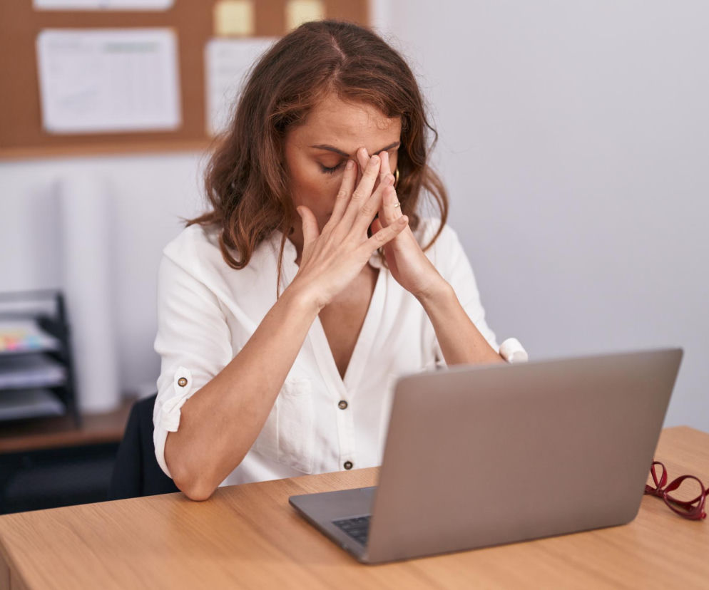 Kobieta z zamkniętymi oczami, z dłońmi opartymi na skroniach, siedzi przy laptopie na drewnianym biurku, zmęczona lub zmagająca się z brakiem koncentracji. W tle widać tablicę korkową i organizer na dokumenty. Artykuł na Poradnik Zdrowie wyjaśnia, jak śniadanie wpływa na popołudniową produktywność.