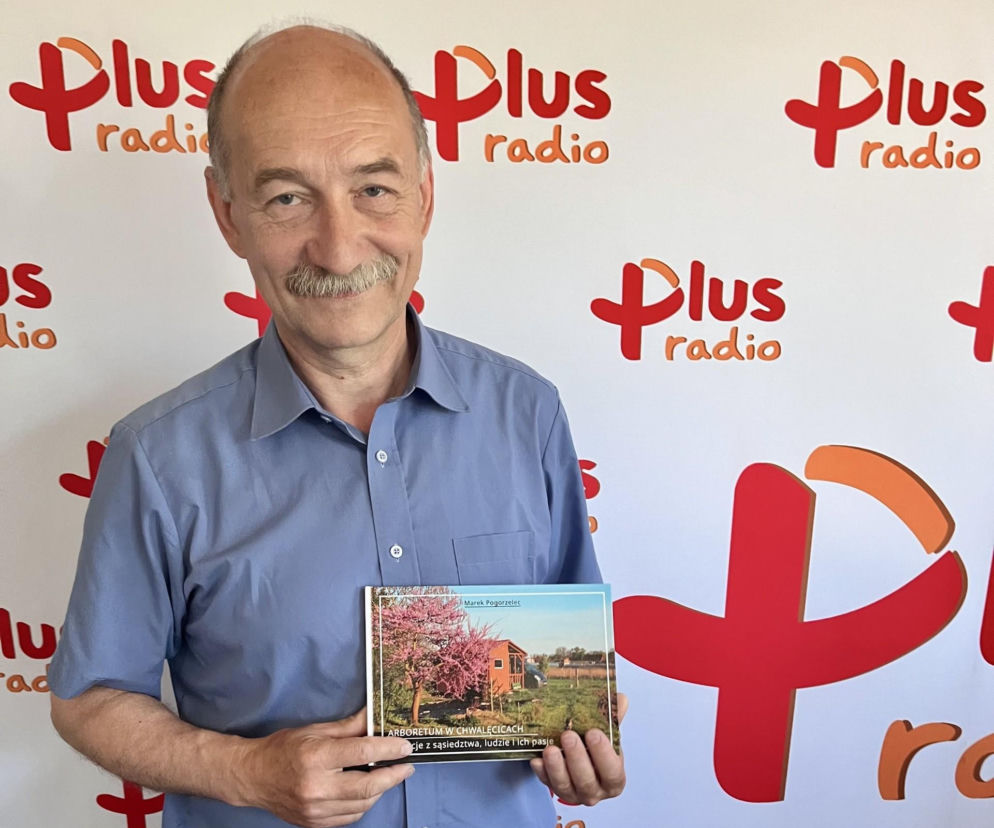 Mężczyzna, Marek Pogorzelec, w błękitnej koszuli, z wąsami, trzyma w dłoniach książkę „Arboretum w Chwalęcicach. Atrakcje z sąsiedztwa, ludzie i ich pasje”, stojąc przed ścianą z logo „Plus Radio”. Więcej o działalności Pogorzelca i jego książce przeczytasz na naszym portalu.