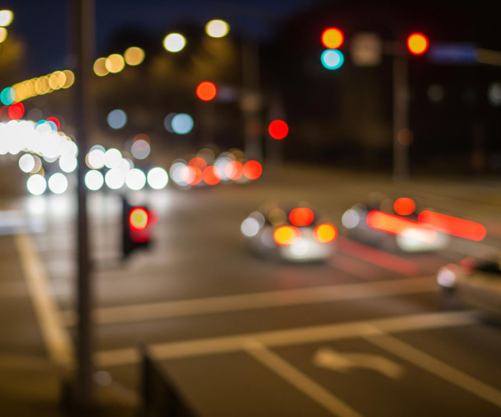 Rozmyty widok przedstawia nocny ruch uliczny z dominującymi światłami w różnych kolorach. Na pierwszym planie po prawej stronie widać rozmazane czerwone smugi świateł tylnych samochodów oraz białe, okrągłe światła przednie pojazdów. W centralnej części obrazu znajduje się ciemna nawierzchnia jezdni z niewyraźnymi białymi liniami pasów ruchu i oznaczeniami poziomymi. W lewej górnej części widoczne są żółte i białe okrągłe plamy świetlne, prawdopodobnie pochodzące z latarni ulicznych lub odległych świateł pojazdów. Na dalszym planie znajdują się sygnalizatory świetlne, z których jeden świeci na zielono, a drugi na czerwono, umieszczone na ciemnych słupach.