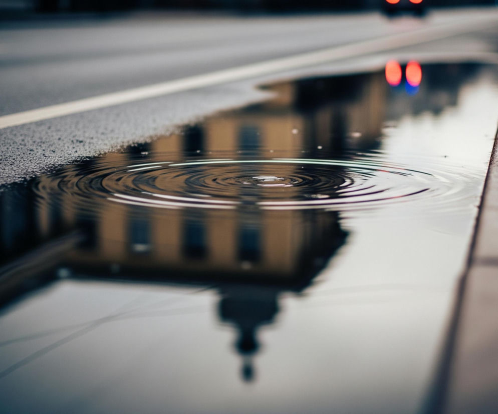 W kałuży na ulicy, położonej wzdłuż krawężnika, odbija się rozmyty obraz żółtawego budynku z ciemnymi oknami. Na powierzchni wody, w centrum kałuży, widoczne są koncentryczne fale. Czarne, mokre pasy asfaltu z białą linią oddzielają kałużę od dalszej części jezdni, gdzie w oddali, poza ostrością, widać czerwone światła hamowania samochodu. Całość jest ciemna i nastrojowa, z dominującymi odcieniami szarości i brązu.