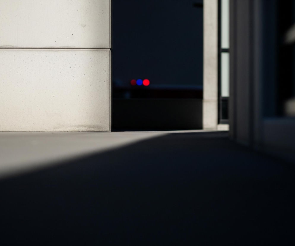 Na pierwszym planie, w lewej części kadru, znajduje się betonowa ściana w odcieniu jasnej szarości, z widocznymi drobnymi nieregularnościami i poziomą fugą dzielącą ją na dwie części. Prawa krawędź ściany styka się z ciemną przestrzenią, z której wyłaniają się dwa rozmyte, intensywnie kolorowe światła: jedno czerwone i jedno niebieskie. Na dole kadru rozciąga się ciemny cień, który ukośnie przecina dolną powierzchnię, przechodząc od jasnoszarego do niemal czarnego. W tle, po prawej stronie, widać rozmyte elementy konstrukcyjne, tworzące pionowe i poziome linie, sugerujące fragmenty okna lub balustrady.