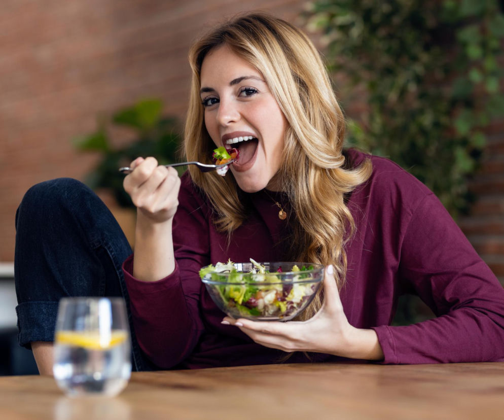 Uśmiechnięta młoda kobieta z długimi, blond włosami je zdrową sałatkę z przezroczystej miski, trzymając widelec blisko ust. Na drewnianym stole obok niej stoi szklanka wody z cytryną. Tło stanowi ceglana ściana i zielona roślina. Więcej o zdrowym odżywianiu przeczytasz na Poradnik Zdrowie.