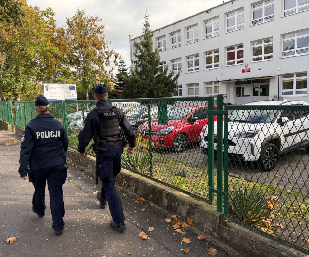 Dwoje policjantów idzie chodnikiem wzdłuż ogrodzenia szkoły. O patrolach w Toruniu przeczytasz na Eska Toruń.