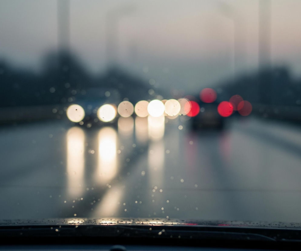 Początek deski rozdzielczej samochodu z widocznymi kroplami wody na przedniej szybie, która jest częściowo zamglona, tworzy dolną część kadru. Na mokrej drodze odbija się światło z rzędu rozmytych samochodów jadących w przeciwnym kierunku, z których pierwsze mają jasne, okrągłe reflektory, a dalsze, w oddali, ukazują czerwone światła tylne. Całość utrzymana jest w chłodnych, szaro-niebieskich barwach z ciepłymi akcentami świateł.