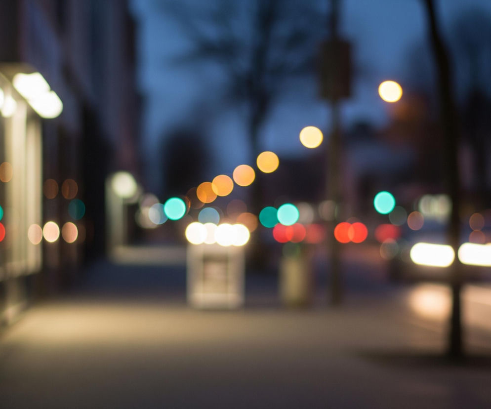 Rozmyte ujęcie ulicy miejskiej nocą, z widocznym chodnikiem po lewej stronie, oświetlonym przez jasne, białe światło pochodzące z fasady budynku lub latarni. W dali rozciągają się liczne, kolorowe punkty świetlne o kształcie okręgów, głównie pomarańczowe, czerwone i turkusowe, symbolizujące światła uliczne i samochodowe. Niebo w górnej części kadru ma ciemnoniebieski odcień, a po prawej stronie majaczy rozmazana sylwetka drzewa oraz smugowe światła pojazdów.