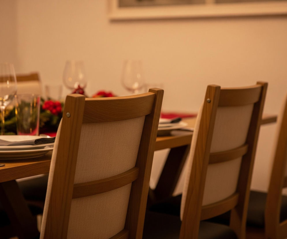 Trzy drewniane krzesła z jasnymi, materiałowymi oparciami stoją przy prostokątnym, drewnianym stole, na którym ułożone są białe talerze z szarymi serwetkami. Na stole widoczny jest również świąteczny stroik z zielonymi gałązkami, czerwonymi owocami i złotymi ozdobami, a obok niego pali się jasna świeca. W tle, częściowo niewyraźne, stoją kieliszki do wina, a pomieszczenie oświetlone jest ciepłym, żółtym światłem.