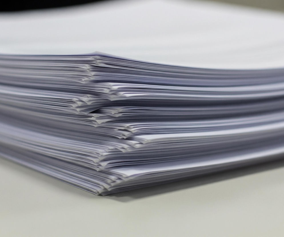 Na pierwszym planie, w lewym dolnym rogu, znajduje się duża sterta białych kartek papieru, ułożonych jedna na drugiej, tworząc nieregularny, pofalowany kształt. Kartki są różnej grubości i mają lekko zagięte krawędzie, co sprawia wrażenie, że nie są idealnie równe. Kolor papieru jest biały, z odcieniami szarości widocznymi na bocznych krawędziach i w cieniach między kartkami. Tło jest rozmyte, utrzymane w jasnych, neutralnych barwach, prawdopodobnie ukazujące biurko lub inną płaską powierzchnię.