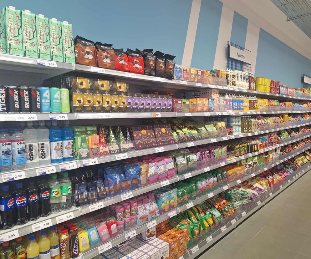 Sklepowe półki pełne napojów i słodyczy z widocznymi cenami. O drożejących produktach przeczytasz na SE Superbiz.