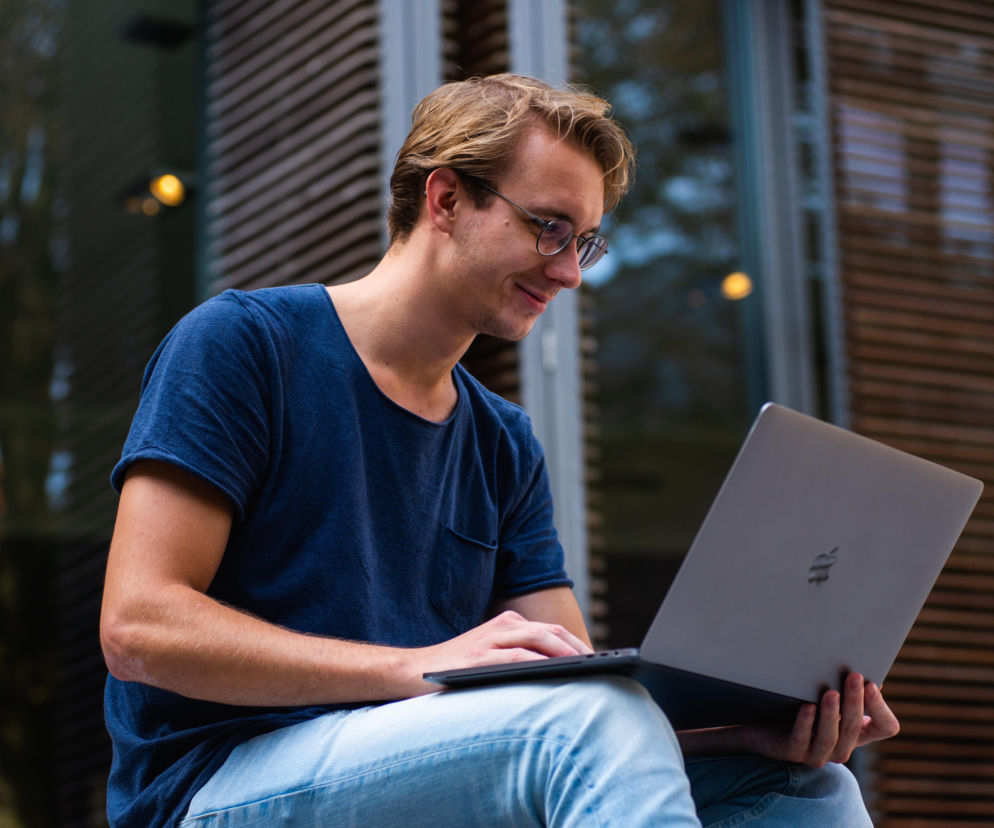 Uśmiechnięty student w okularach korzysta z laptopa na zewnątrz. O popularnych kierunkach studiów przeczytasz na SE.