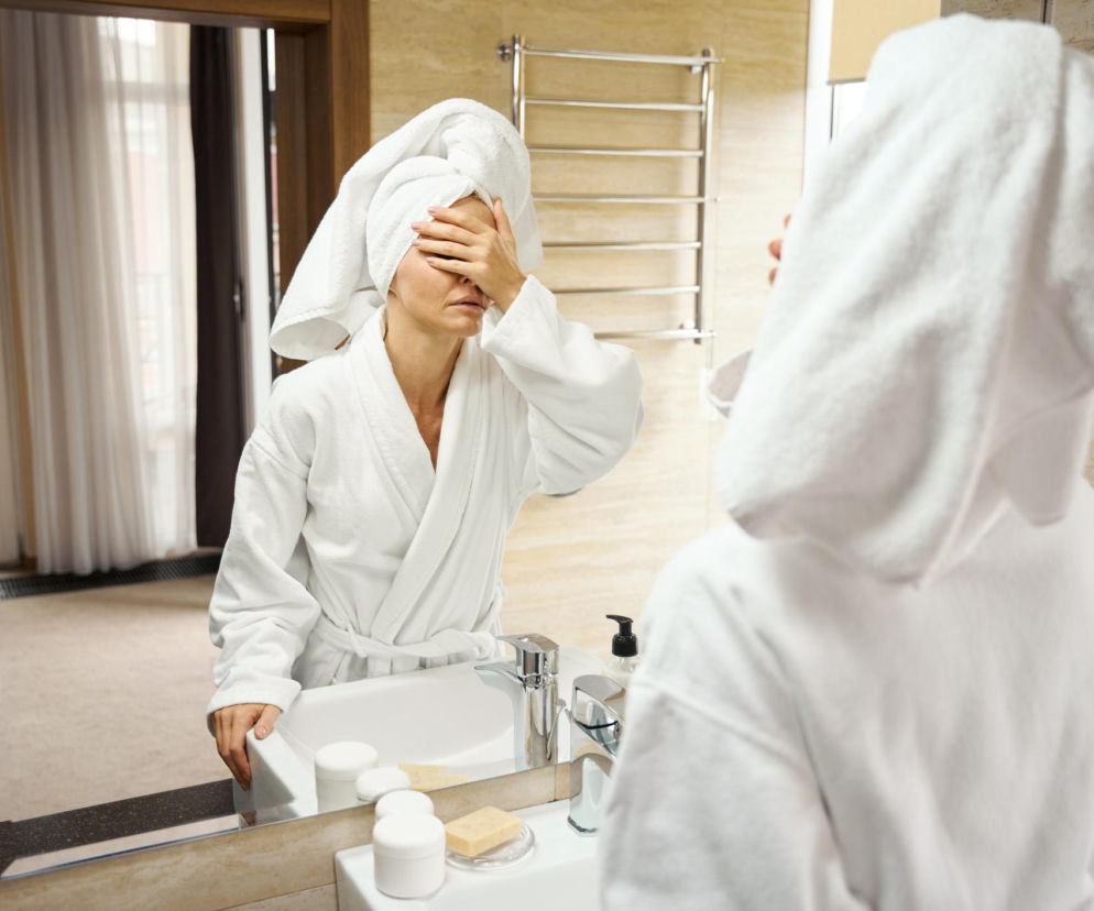 Kobieta w białym szlafroku i z ręcznikiem na głowie trzyma rękę na czole, patrząc w lustro po kąpieli, co może wskazywać na zawroty głowy. Dowiedz się więcej o przyczynach i profilaktyce tego zjawiska na portalu Poradnik Zdrowie.