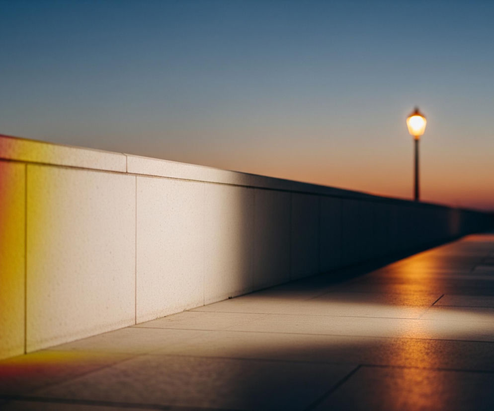 Płytka, jasnobeżowa ściana biegnie ukośnie od lewego dolnego rogu w prawą górę, dzieląc obraz. Na jej powierzchni, po lewej stronie, rozciąga się pionowy pas kolorów tęczy: od ciemnego fioletu, przez niebieski, zielony, żółty, pomarańczowy, aż po czerwień. Jasne, pomarańczowe światło rzuca się również ukośnie na podłogę z jasnych, kwadratowych płytek, tworząc kontrast z ciemniejszymi obszarami podłogi po prawej. W oddali, na horyzoncie, widoczna jest rozmyta sylwetka słupa z pojedynczą, jasną lampą uliczną, a niebo zmienia barwy od błękitu u góry do delikatnego pomarańczu nad horyzontem.