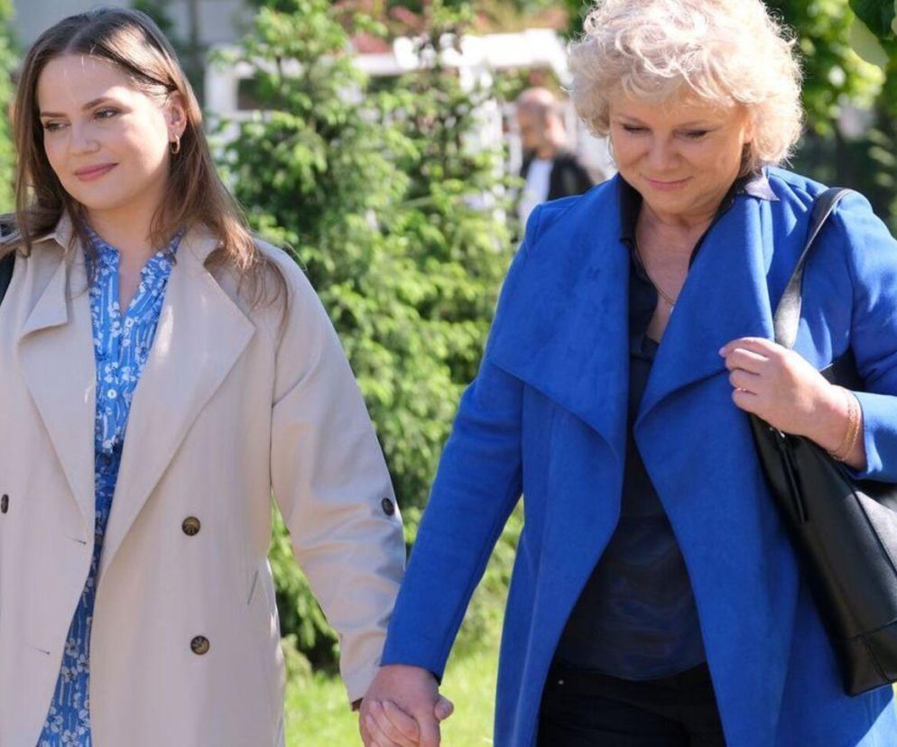 Anita (Melania Grzesiewicz) i Anna (Dorota Kamińska) z M jak miłość trzymające się za ręce. Anna w niebieskim płaszczu i czarnej torebce, Anita w beżowym trenczu i wzorzystej koszuli. Obie idą po ścieżce, w tle widać zieleń i fragmenty budynku. Więcej o tym na Super Seriale.