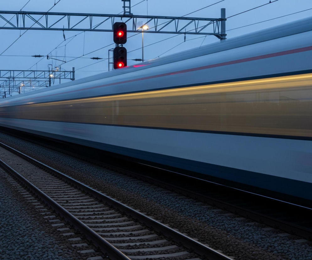 Na pierwszym planie, z lewej strony, widoczne są dwa czerwone światła semafora umieszczonego na słupie, które jaśnieją w mroku. Na dalszym planie, po prawej stronie, rozciąga się pociąg o metalicznym, srebrzysto-niebieskim kolorze z poziomym czerwonym pasem, który przemieszcza się z dużą prędkością, co jest widoczne dzięki rozmytemu efektowi ruchu. Z dołu, wzdłuż lewej krawędzi pociągu, biegną dwa tory kolejowe, a ich podłoże pokryte jest drobnym żwirem; po prawej stronie torów widoczne są fragmenty roślinności oraz betonowy krawężnik. Nad pociągiem rozciąga się konstrukcja trakcyjna z licznymi kablami i masztami, a całość ujęta jest pod niebem w odcieniach szarości i błękitu o zmierzchowej porze.