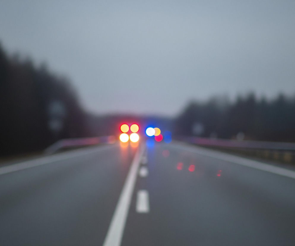 Rozmazaną perspektywę drogi szybkiego ruchu w niekorzystnych warunkach pogodowych, prawdopodobnie zmierzchu lub wczesnego poranka, z widoczną mgłą lub zamgleniem. Centralną część zajmuje szara, asfaltowa jezdnia z wyraźną, białą, ciągłą linią pośrodku oraz przerywanymi liniami po prawej stronie. W oddali, na horyzoncie, widoczne są rozświetlone światła samochodów: cztery pomarańczowo-żółte punkty w górnej części oraz niebieskie i czerwone punkty świetlne po ich bokach. Po obu stronach drogi, w tle, zarysowują się ciemne sylwetki drzew lub lasu, również niewyraźne.