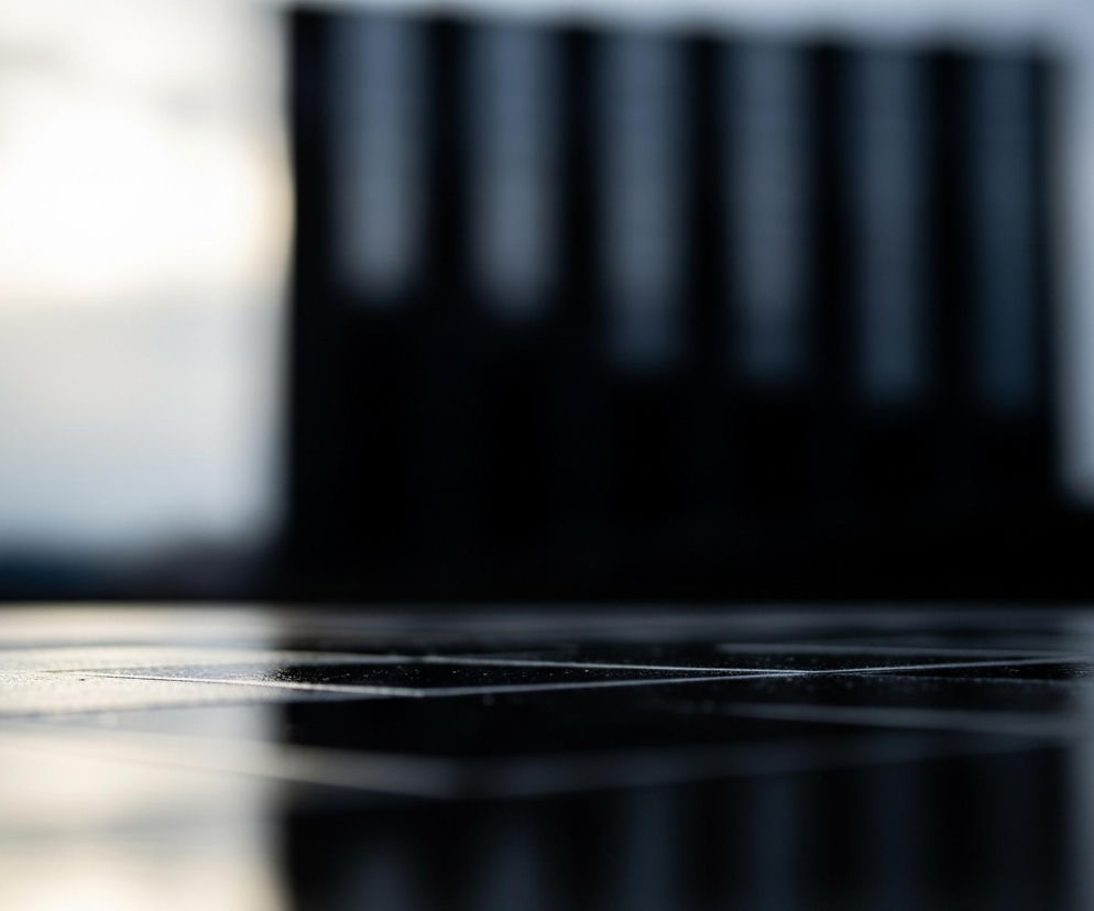 Pierwszy plan zajmuje powierzchnia składająca się z kwadratowych płytek w ciemnych odcieniach szarości i czerni, z delikatnymi, jasnymi refleksami i widocznymi drobnymi kropelkami lub zabrudzeniami. Płytki są błyszczące, odbijając światło i niewyraźne kształty. Tło jest rozmyte, dominują w nim pionowe, ciemne pasy, które w środkowej części obrazu są prawie czarne, a po bokach jaśniejsze, wpadające w odcień błękitu i szarości. Lewa strona tła charakteryzuje się jaśniejszym, rozbielonym światłem, podczas gdy prawa strona jest bardziej jednolita w odcieniach szarości.