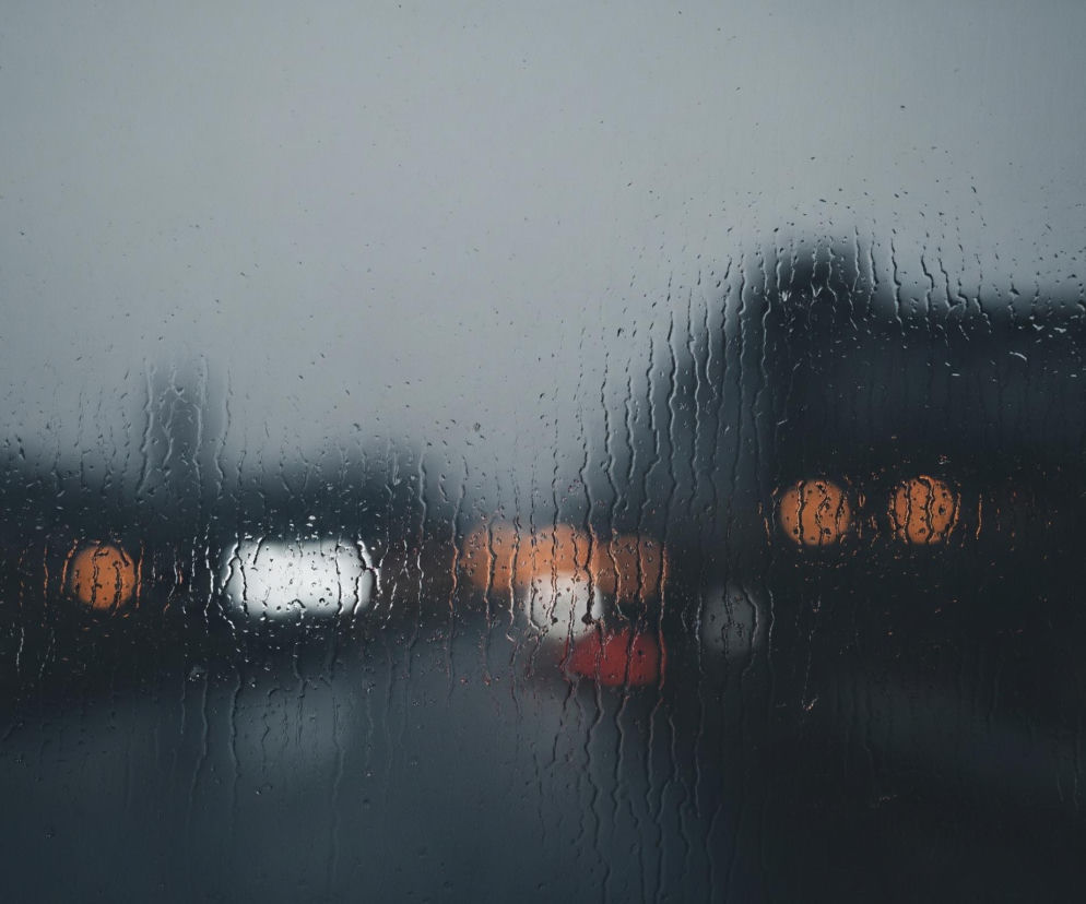 Przednia szyba, pokryta licznymi kroplami deszczu i smugami wody, tworzy pierwszy plan obrazu. Woda zniekształca i rozmywa tło, które przedstawia nocny widok miasta z rozmytymi światłami pojazdów. W oddali widoczne są jasne, białe reflektory, pomarańczowe światła uliczne oraz czerwone światła tylne. Ogólna kolorystyka jest ciemna i chłodna, z dominującymi odcieniami szarości, błękitu i czerni, przełamanymi ciepłymi, rozmytymi punktami światła.