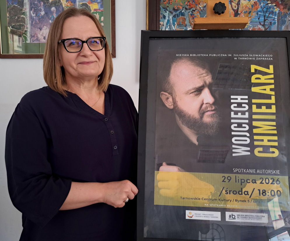 Kobieta w okularach stoi obok plakatu zapraszającego na spotkanie z Wojciechem Chmielarzem. Szczegóły na Eska Tarnów.