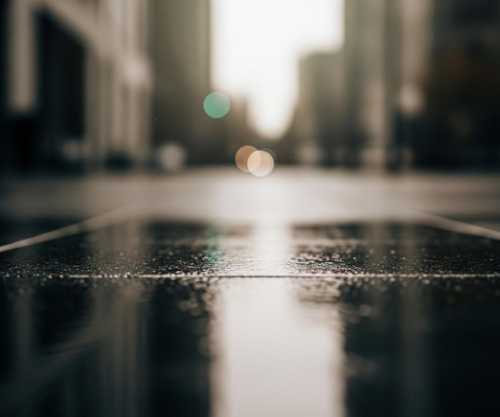 Na pierwszym planie widoczna jest mokra, ciemna nawierzchnia z drobnymi kropelkami wody odbijającymi światło. Biegną przez nią dwie jasne, równoległe linie, które zbiegają się ku górze obrazu, tworząc wrażenie głębi. Tło jest rozmyte i nieostre, ukazując niewyraźne kształty budynków oraz kilka okrągłych, kolorowych plam światła, przypominających bokeh. Dominują ciemne, stonowane barwy, a jedynie górna część obrazu rozjaśniona jest przez blask, prawdopodobnie słońca lub innych źródeł światła.