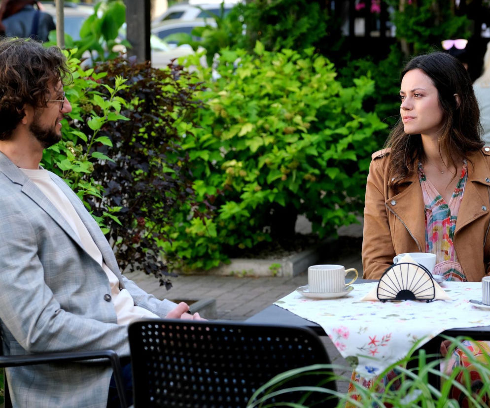 Aktor Mateusz Mosiewicz (Mariusz) i aktorka Paulina Lasota (Kasia) siedzą przy stoliku w restauracyjnym ogródku, patrząc na siebie, z filiżankami kawy przed nimi. To kadr z serialu M jak miłość, o którym więcej przeczytasz na stronie Super Seriale.