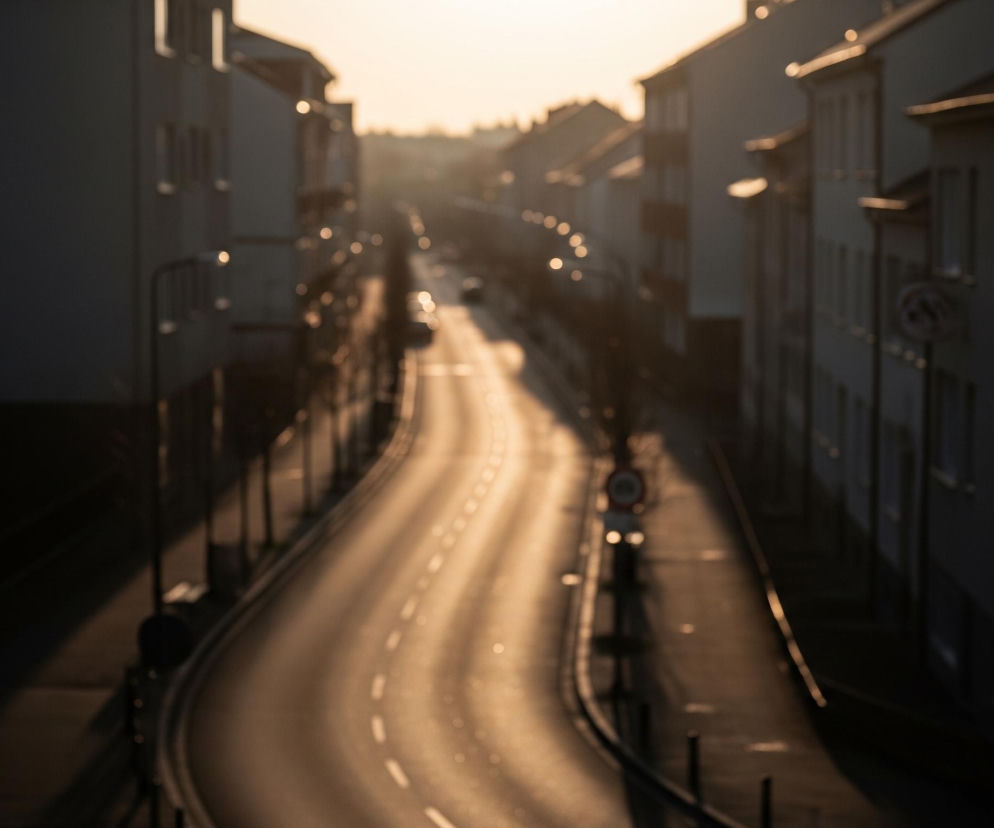 Długa, falista ulica biegnie przez centrum kadru, oświetlona ciepłym, złotym światłem zachodzącego słońca, tworząc na asfalcie podłużne, jasne refleksy i rozmyte świetlne kręgi. Po obu stronach ulicy stoją rzędy wielopiętrowych budynków o ciemnych fasadach, z pojedynczymi, rozmazanymi oknami, niektóre z nich posiadają balkony. W oddali, na końcu ulicy, widać intensywny blask słońca, który rozjaśnia niebo i budynki, tworząc zamgloną perspektywę.