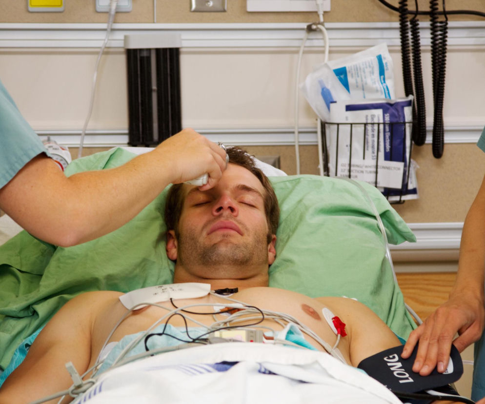 Mężczyzna z elektrodami EKG na klatce piersiowej leży w łóżku szpitalnym, a pielęgniarz mierzy mu temperaturę na czole. Zdjęcie ilustruje, jak infekcja, np. grypa, może zwiększyć ryzyko zawału serca, o czym pisze Poradnik Zdrowie.