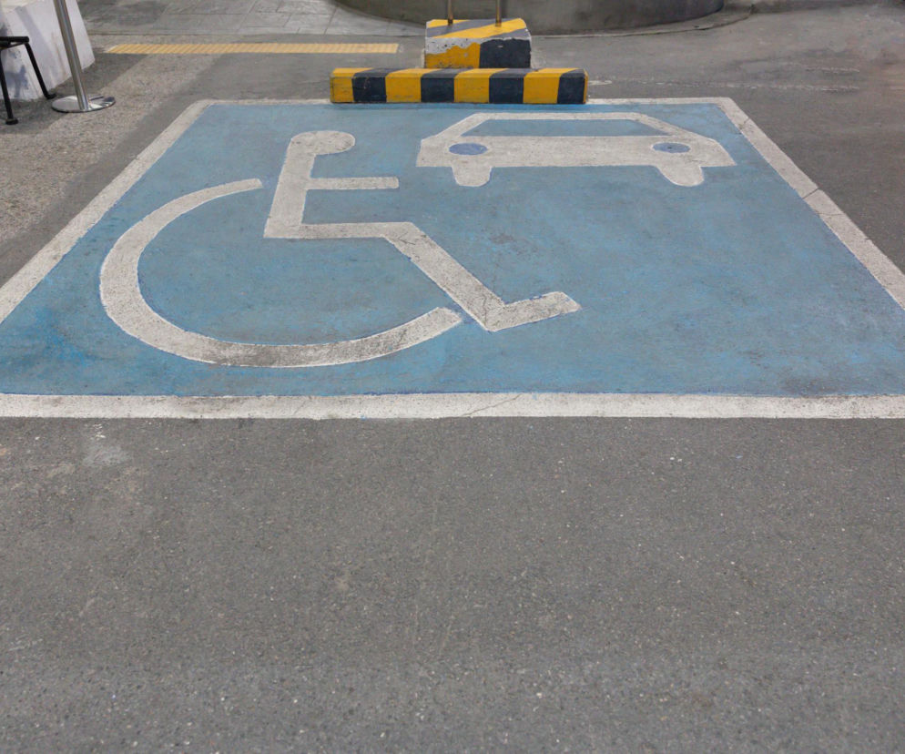 Niebieskie miejsce parkingowe dla osób z niepełnosprawnościami, oznaczone białym symbolem wózka inwalidzkiego i samochodu, na szarym asfalcie. Za nim widoczne czarno-żółte blokady, symbolizujące problem sprzedaży miejsc dla niepełnosprawnych osobom zdrowym, o którym przeczytasz na Poradnik Zdrowie.
