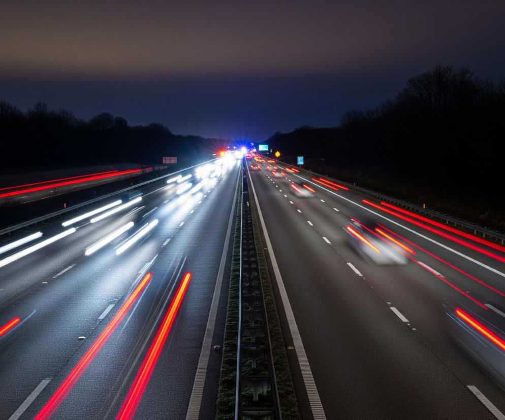 Autostradę nocą z widocznymi smugami świateł pojazdów. Dwie jezdnie rozdzielone są pasem zieleni i barierkami, a w tle widać ciemny, rozmyty las i pas ciemnego nieba. Na lewej jezdni widoczne są intensywne białe smugi świateł mijania oraz czerwone smugi świateł tylnych, a na prawej jezdni przeważają czerwone smugi świateł tylnych. W oddali na prawej jezdni świecą niebieskie i pomarańczowe światła alarmowe.