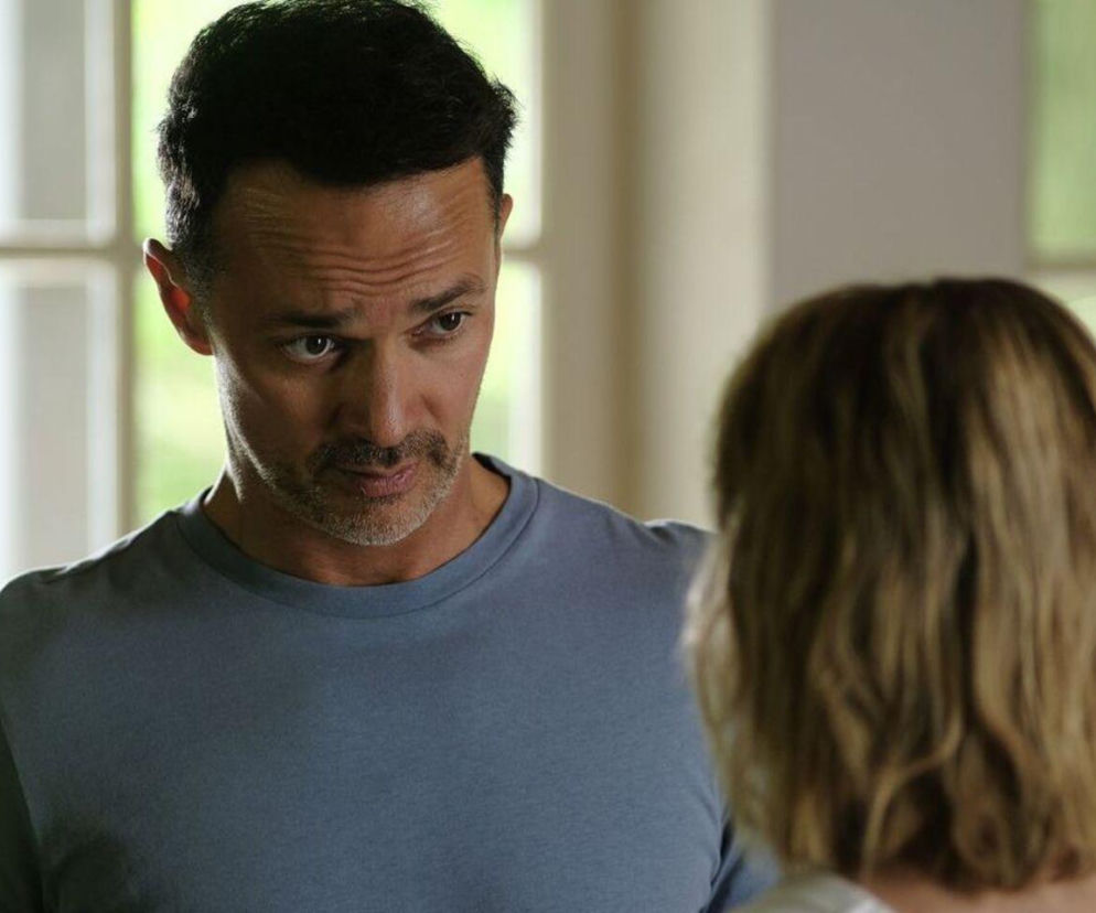 Mariusz (Rafał Cieszyński) w jasnoniebieskiej koszulce z krótkim zarostem, intensywnie spoglądający na tył głowy Janki (Oliwia Drożdżyk), której blond włosy są lekko falowane. W tle widoczne okno i zieleń, co nadaje scenie naturalne oświetlenie. Zdjęcie przedstawia fragment fabuły serialu „Barwy szczęścia”, o którym więcej przeczytasz na portalu Super Seriale.